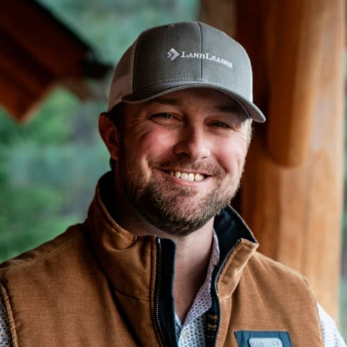A man with a beard wearing a gray "LandLeader" cap and brown vest smiles at the camera in a rustic, outdoor setting.