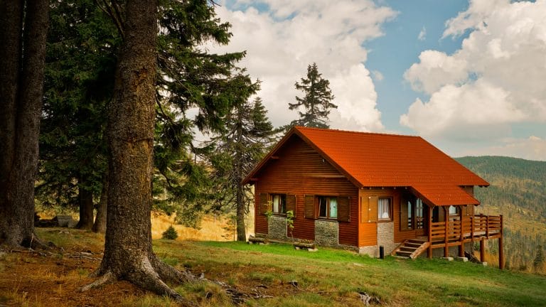 A wooden cabin with a red roof stands on a grassy hillside, surrounded by trees, with mountains and a partly cloudy sky in the background.